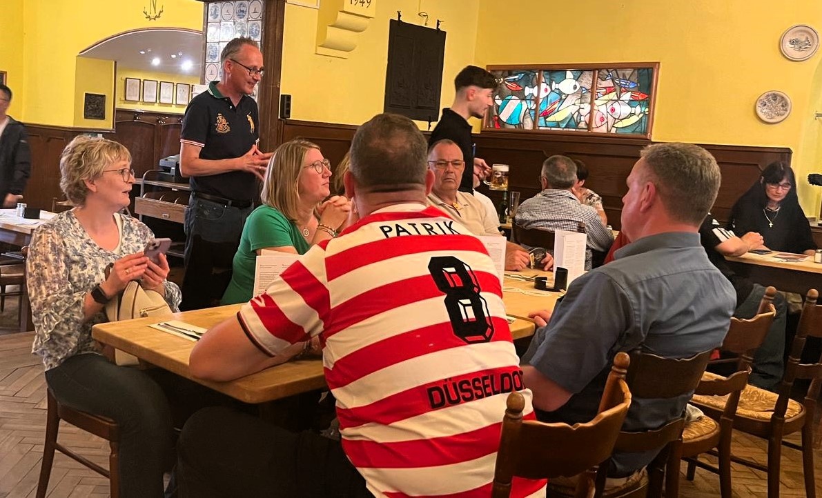 [{"clang_id":1,"value":"Gruppe Menschen sitzt an Tischen in einem Restaurant. Ein Mann trägt ein rotes und weiß gestreiftes Fußballtrikot mit der Aufschrift \"PATRIK\" und \"DÜSSELDORF\"."}]