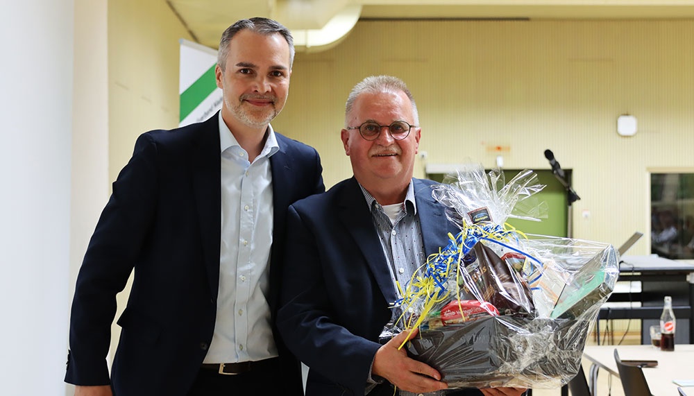 [{"clang_id":1,"value":"Zwei Männer im Anzug stehen nebeneinander und lächeln. Der rechte Mann hält einen Geschenkkorb mit Dekoration. Im Hintergrund ein heller Innenraum."}]
