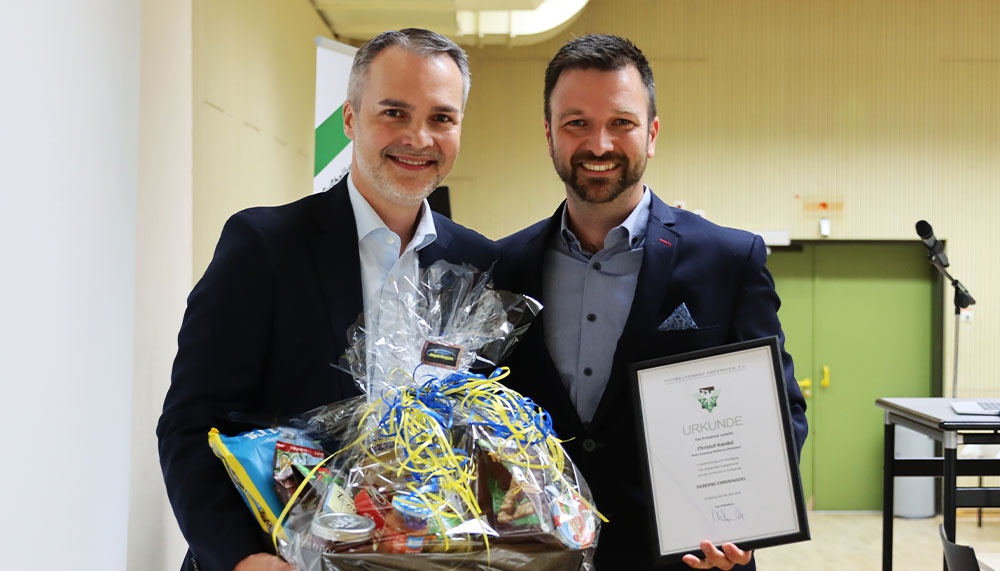 [{"clang_id":1,"value":"Zwei lächelnde Männer in Anzug und Hemd halten jeweils einen Geschenkkorb bzw. eine Urkunde in der Hand. Im Hintergrund eine helle Wand und eine Tafel."}]