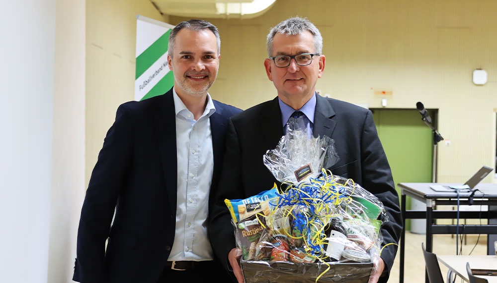 [{"clang_id":1,"value":"Zwei Männer in Anzug und Hemd stehen nebeneinander und halten einen Geschenkkorb mit Lebensmitteln. Im Hintergrund eine Turnhalle."}]