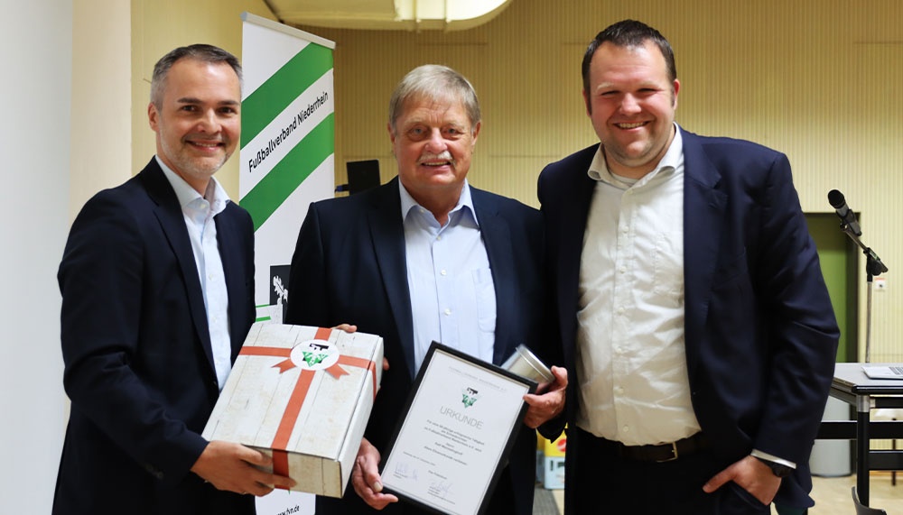 [{"clang_id":1,"value":"Drei Männer in Anzügen stehen nebeneinander und halten Geschenke bzw. eine Urkunde. Im Hintergrund ein Banner mit Schriftzug."}]