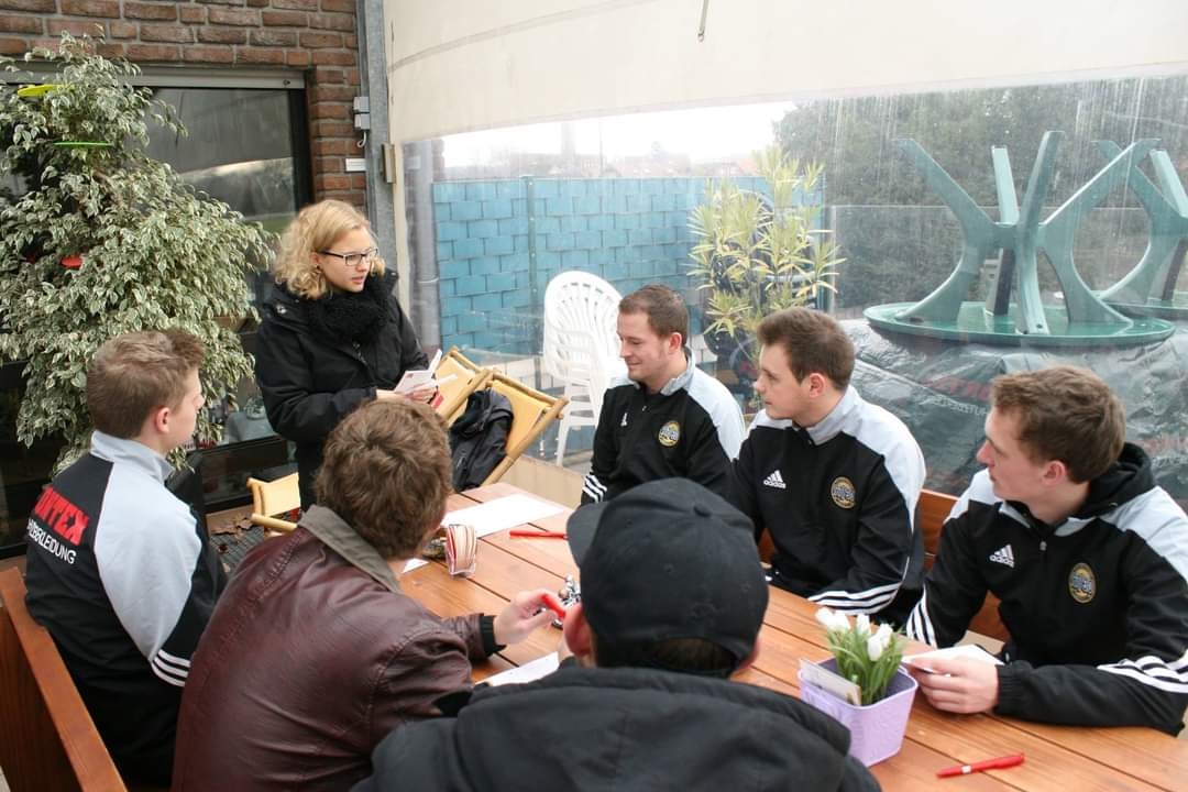 [{"clang_id":1,"value":"Gruppe junger Männer sitzt an einem Tisch im Freien, eine Frau steht und erklärt etwas. Im Hintergrund ein Garten mit Pflanzen und Skulptur."}]