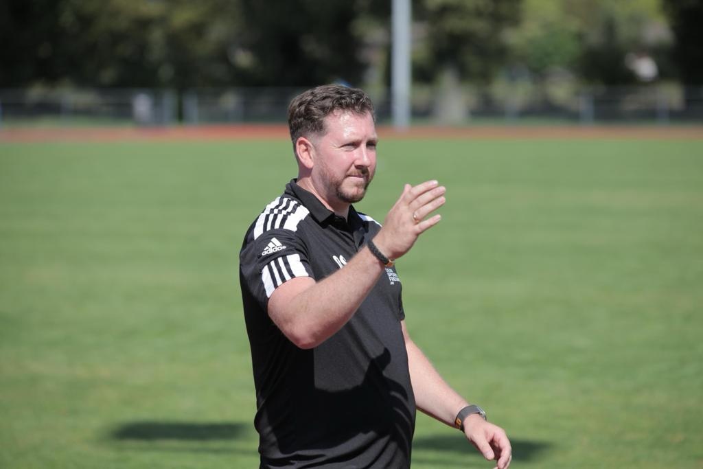 [{"clang_id":1,"value":"Mann mit rotem Bart, schwarzer Sportbekleidung und Adidas-Logo applaudiert auf einem grünen Rasenplatz. Im Hintergrund Bäume und Sportstadion."}]