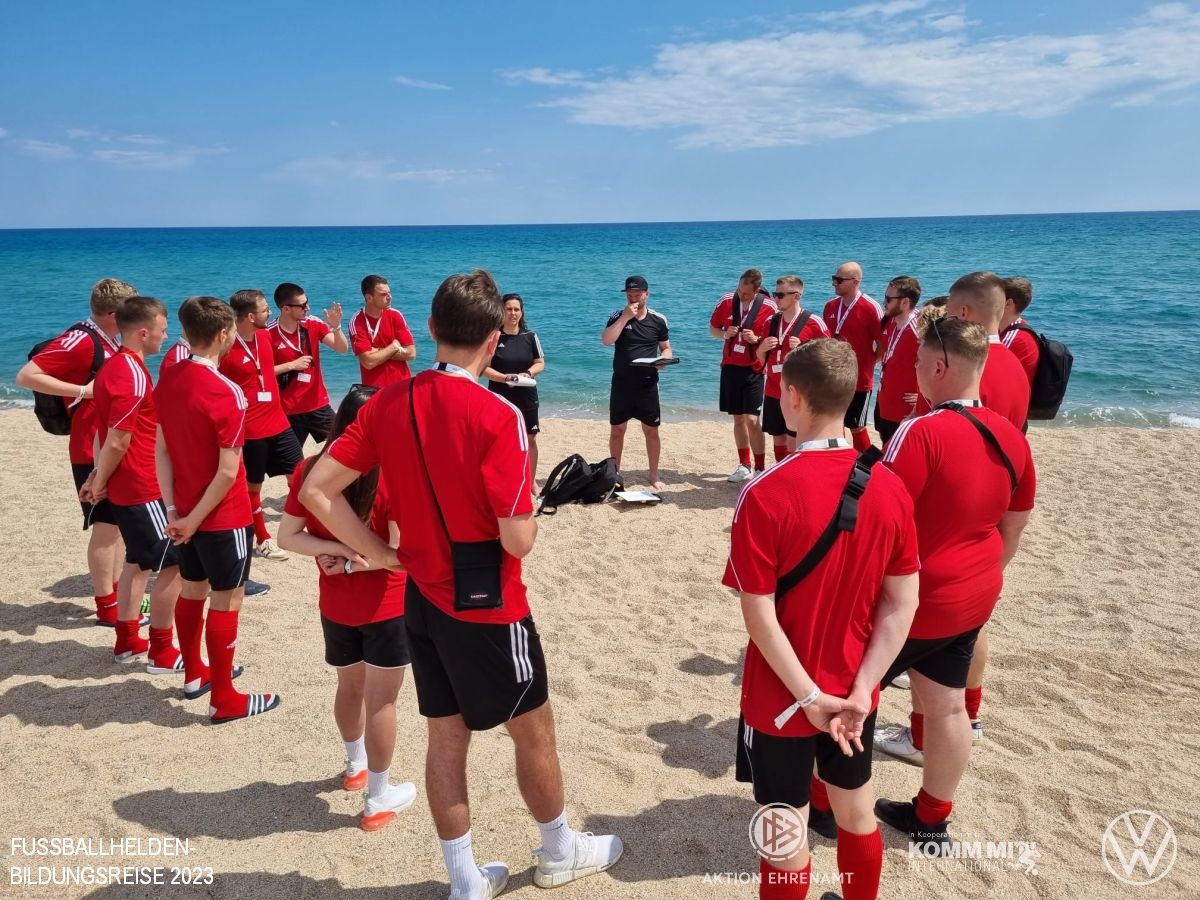 [{"clang_id":1,"value":"Fußballteam in rot-schwarzen Trikots am Strand. Trainer steht vor der Gruppe, Blick auf das Meer im Hintergrund. Logos am unteren Rand."}]