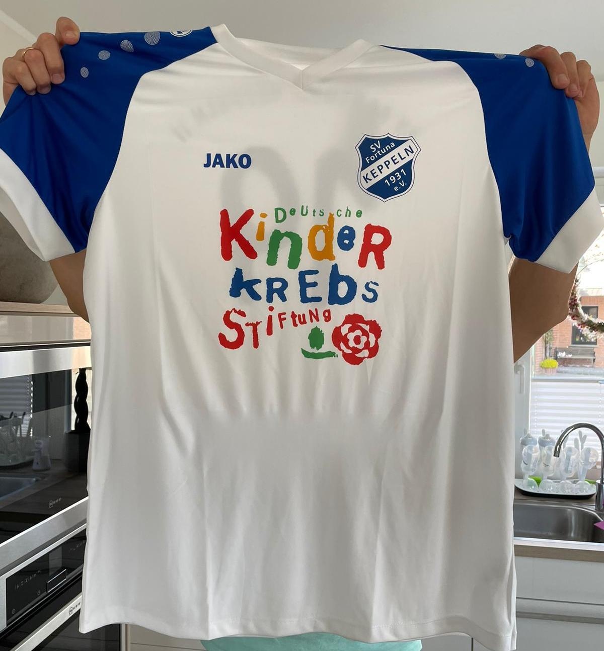 [{"clang_id":1,"value":"Blau-weißes Fußballtrikot hängt an einem Kleiderbügel. Auf der Brust steht in bunten Lettern \"Deutsche Kinder Krebs Stiftung\" und ein Rosen-Logo. Im Hintergrund eine Küchenzeile."}]