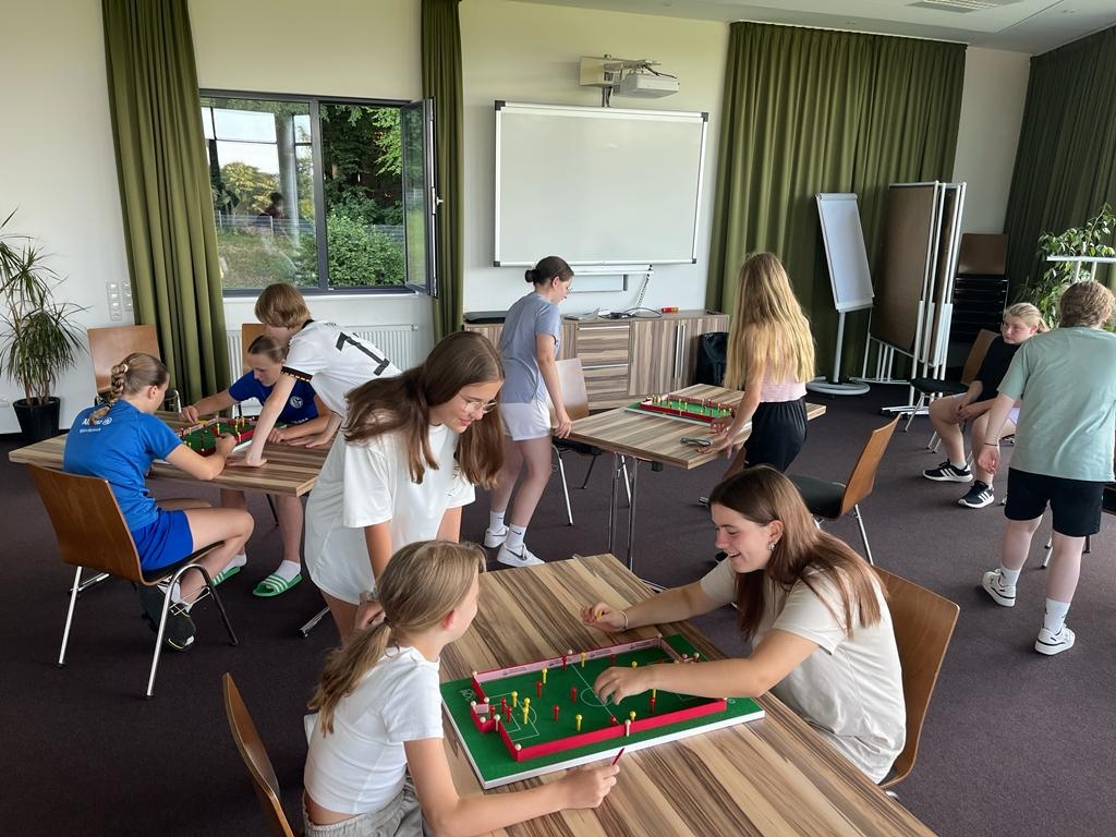 [{"clang_id":1,"value":"Gruppe Jugendlicher spielt Tischfußball in einem hellen Raum mit Fenster und Whiteboard. Einige sitzen an Tischen, andere stehen und schauen zu."}]