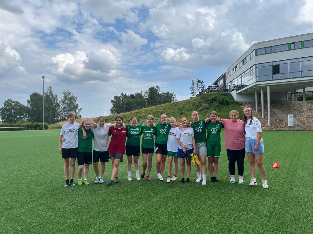 [{"clang_id":1,"value":"Fußballmannschaft mit Mädchen in Sportkleidung steht lächelnd auf grüner Kunstrasenfläche vor einem modernen Gebäude und Bäumen unter bewölktem Himmel."}]