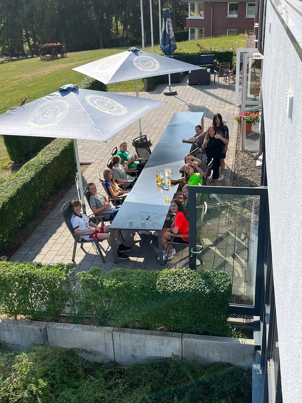 [{"clang_id":1,"value":"Terrasse mit mehreren Personen an langen Tischen unter Sonnenschirmen. Rasenfläche und Gebäude im Hintergrund."}]