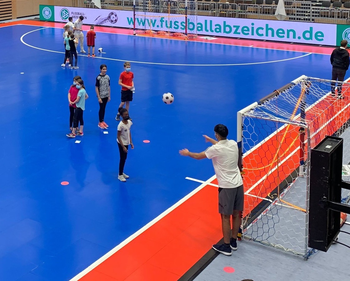 [{"clang_id":1,"value":"Halle mit blauen Boden. Kinder spielen Fußball, einige tragen Masken. Ein Junge schießt auf ein Tor. Im Hintergrund eine Werbeplane."}]