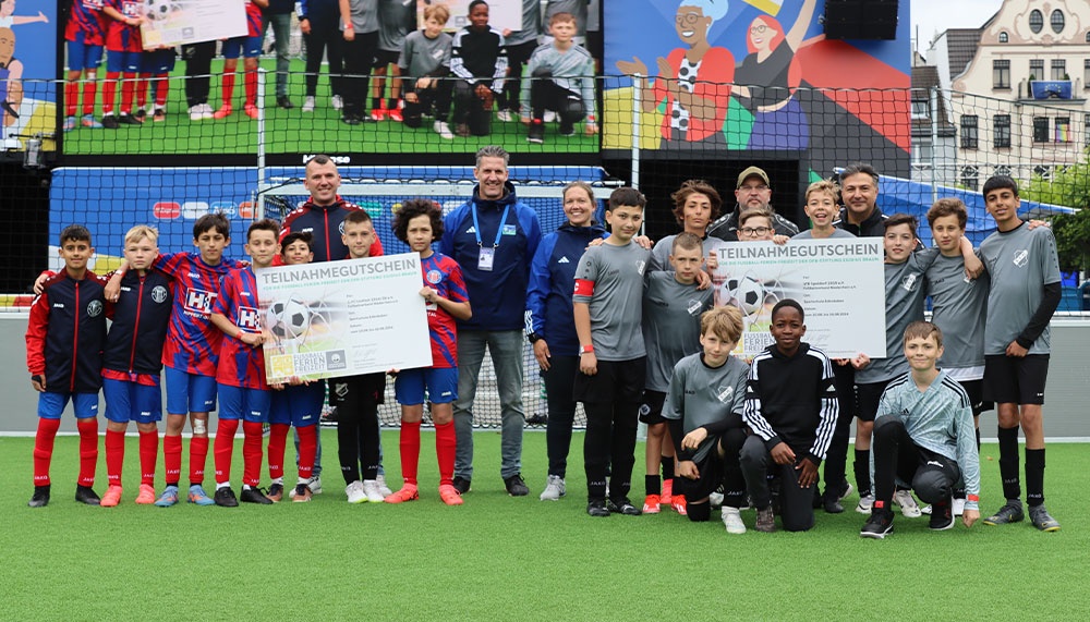 [{"clang_id":1,"value":"Fußballmannschaft mit Betreuern und Kindern auf Kunstrasenfeld. Einige halten Teilnahmezertifikate. Im Hintergrund eine Kulisse mit Gebäuden und Menschen."}]