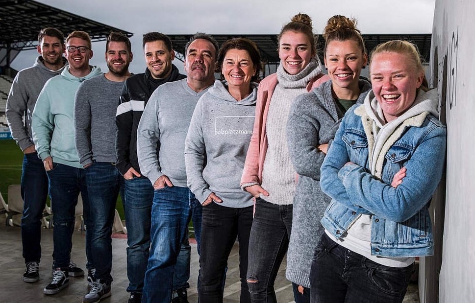 [{"clang_id":1,"value":"Gruppe von neun Personen steht lächelnd nebeneinander vor einer Stadionkulisse. Sie tragen Freizeitkleidung, Jeans und Sweatshirts."}]