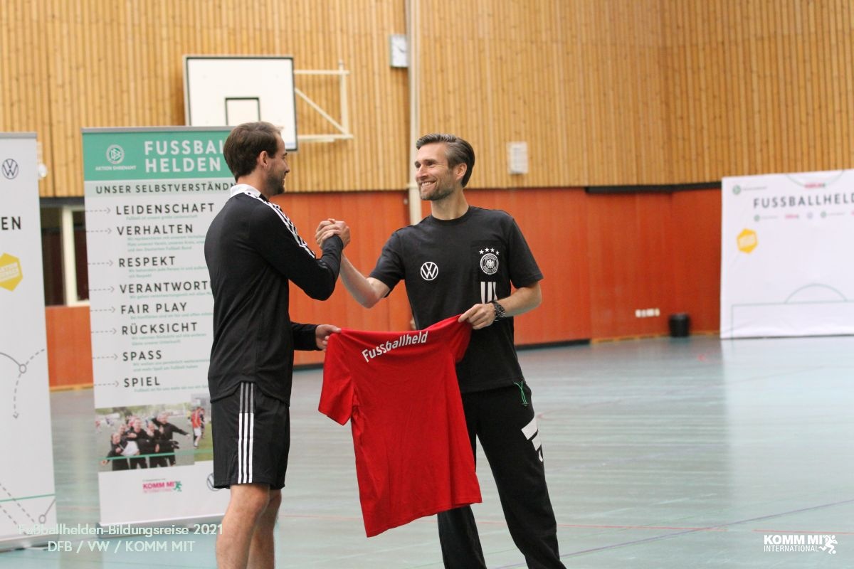 [{"clang_id":1,"value":"Zwei Männer in Sporthalle. Ein Mann übergibt dem anderen ein rotes Fußballtrikot. Im Hintergrund Banner mit Fußball-Werten und Logos."}]