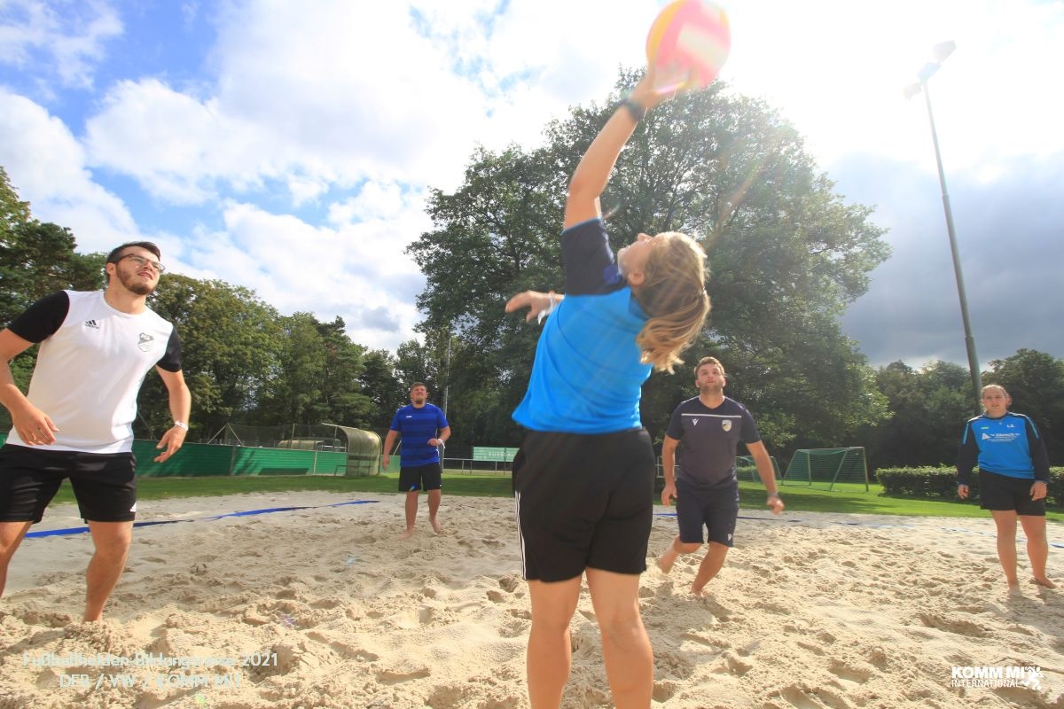[{"clang_id":1,"value":"Volleyballspieler*innen auf Sandfeld unter blauem Himmel. Eine Spielerin wirft Ball. Weitere Spieler*innen stehen bereit. Im Hintergrund Grünflächen und Bäume."}]
