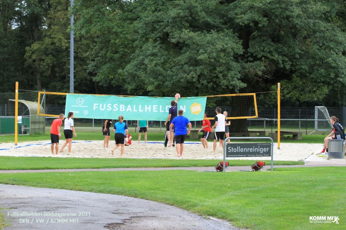 [{"clang_id":1,"value":"Volleyballfeld mit spielenden Personen. Im Hintergrund grüne Bäume und ein Werbebanner mit der Aufschrift \"FUSSBALLHELDEN\". Rasenfläche im Vordergrund."}]