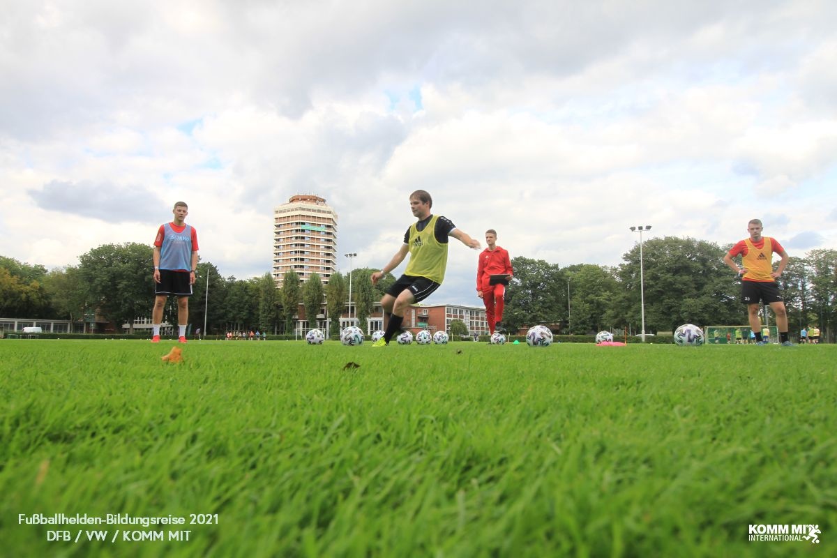 [{"clang_id":1,"value":"Fußballtraining: Fünf junge Männer auf grünem Rasen, einer schießt Ball. Im Hintergrund Gebäude und bewölkter Himmel. Logo \"KOMM MIT INTERNATIONAL\" rechts unten."}]