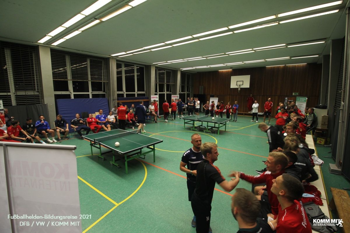 [{"clang_id":1,"value":"Große Sporthalle mit vielen Jugendlichen, die Tischtennis spielen und sich unterhalten. Einige stehen und gestikulieren. Im Hintergrund Fenster und Basketballkorb."}]