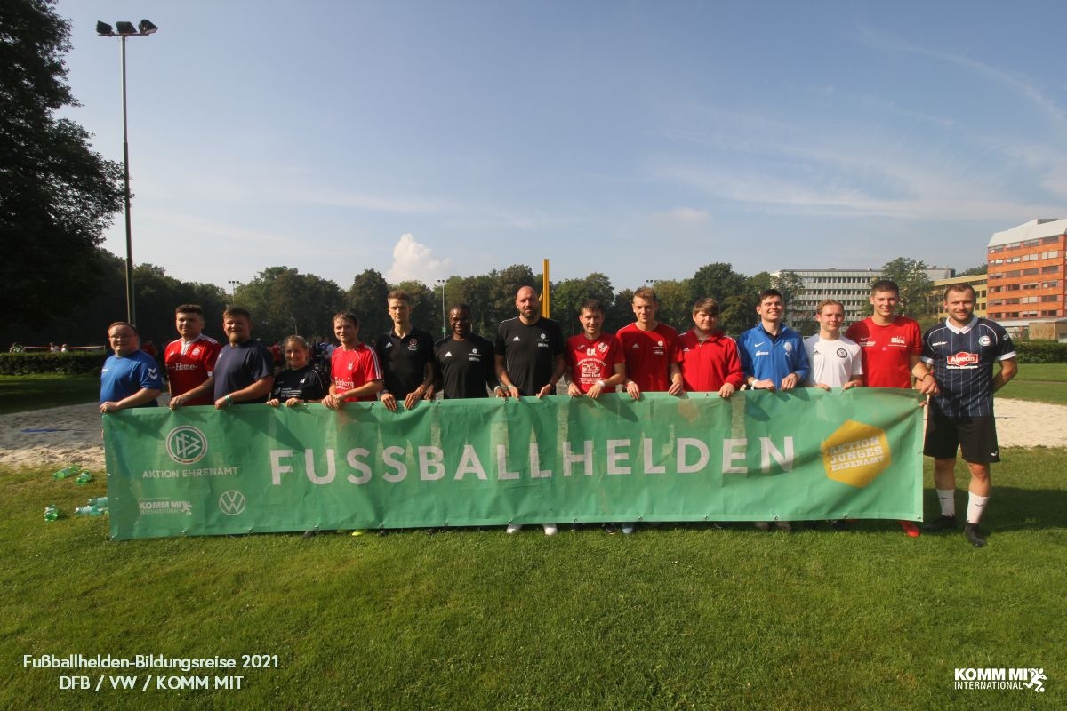 [{"clang_id":1,"value":"Gruppe von rund 15 Personen auf Rasen vor Gebäuden. Sie halten ein grünes Banner mit der Aufschrift \"FUSSBALLHELDEN\" und Logos von VW und Aktion Gesundheit."}]