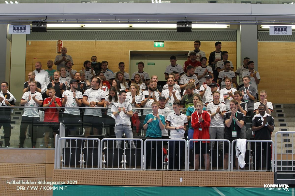 [{"clang_id":1,"value":"Zuschauerreihen in einer Sporthalle. Viele Menschen sitzen und stehen, überwiegend in weißen Oberteilen. Im Vordergrund eine Metallabsperrung."}]