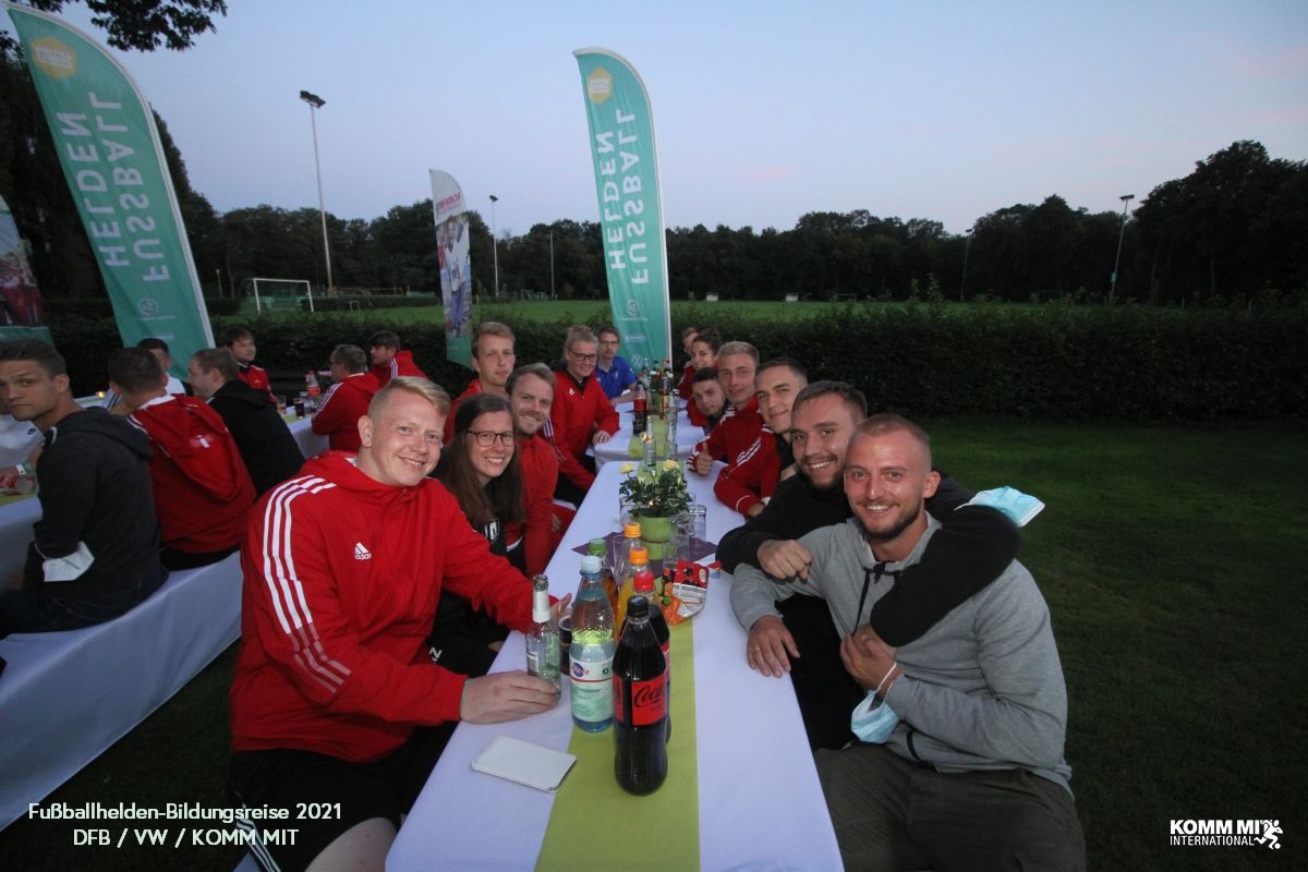 [{"clang_id":1,"value":"Gruppe Menschen sitzt an langen Tischen im Freien, vermutlich nach einem Fußballspiel. Im Hintergrund ein Fußballfeld mit Flutlichtmasten und Bäume."}]