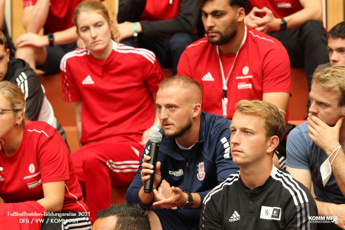 [{"clang_id":1,"value":"Gruppe junger Menschen sitzt in einem Raum. Einige tragen rote Sportshirts, andere schwarze Trainingsanzüge. Ein Mann spricht in ein Mikrofon."}]