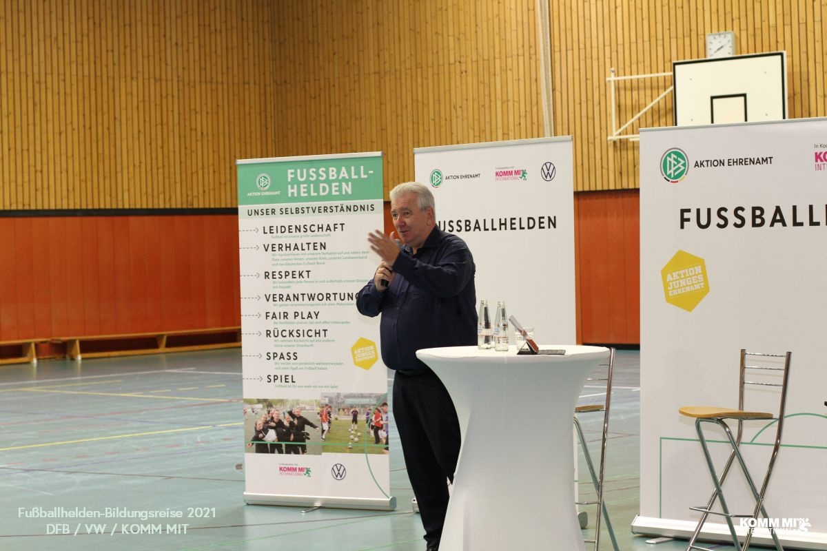 [{"clang_id":1,"value":"Mann am Rednerpult vor Banner mit Fussball-Helden-Logo und Werten wie Leidenschaft, Respekt, Fairplay. Sporthalle im Hintergrund."}]