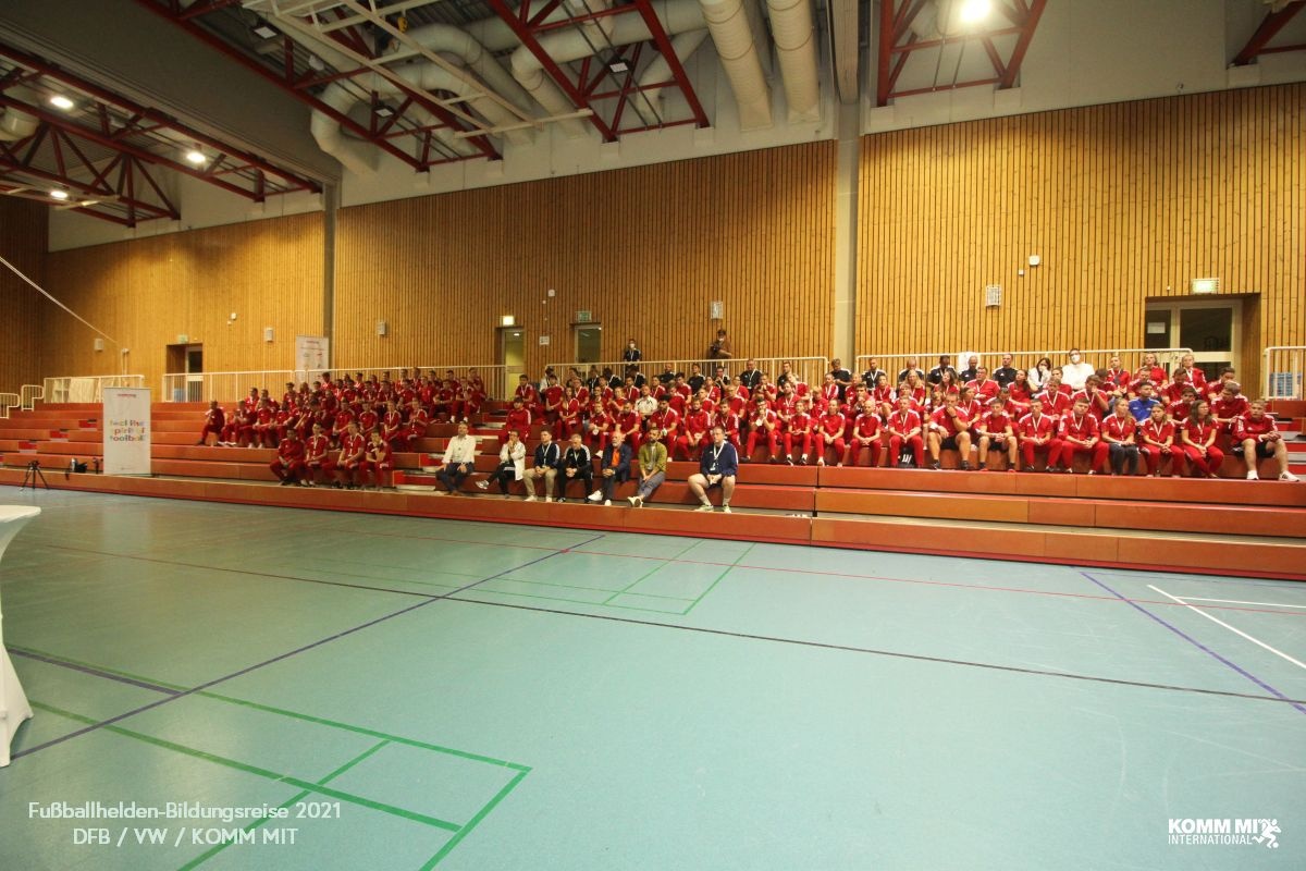 [{"clang_id":1,"value":"Große Sporthalle mit vielen Jugendlichen in roten T-Shirts auf den Zuschauertribünen. Davor einige Betreuer. Auf dem Boden blaue Sportfläche."}]