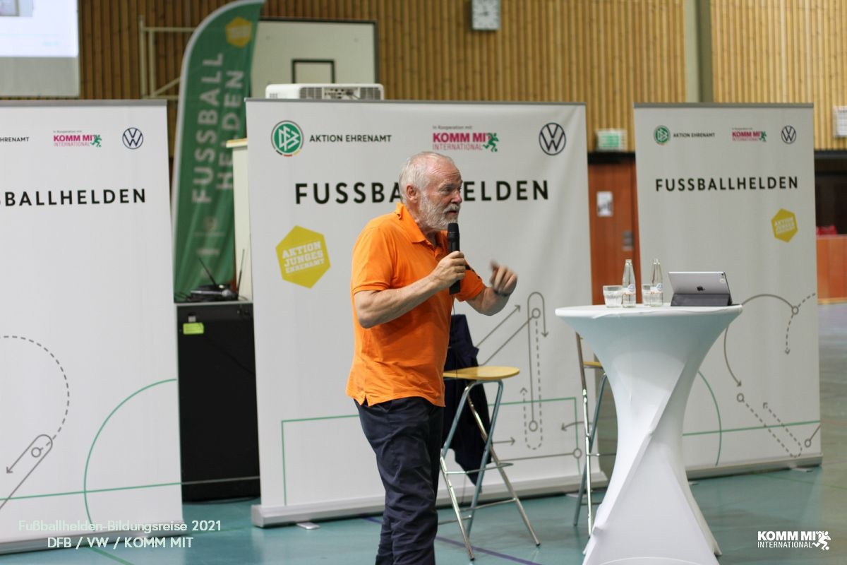 [{"clang_id":1,"value":"Mann in orangefarbenem T-Shirt spricht vor Banner mit Fußballmotiven in einer Sporthalle. Im Hintergrund weitere Banner und Personen."}]