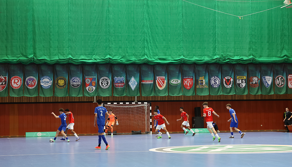 [{"clang_id":1,"value":"Futsalspiel in Sporthalle: Spieler in rot und blau kämpfen um Ball vor Tor. Im Hintergrund grüne Wand mit Fahnen von Sportvereinen."}]