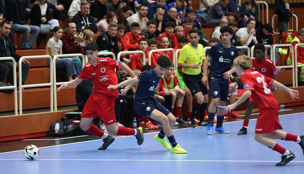 [{"clang_id":1,"value":"Futsalspiel in Halle. Spieler in roten und dunklen Trikots kämpfen um Ball. Publikum auf den Tribünen im Hintergrund. Ein Schiedsrichter ist zu sehen."}]