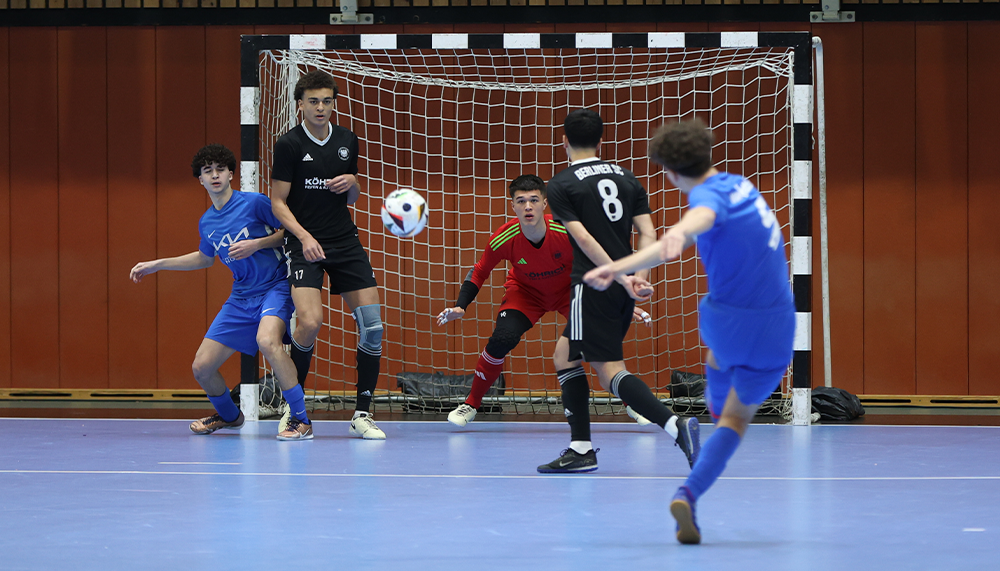 [{"clang_id":1,"value":"Futsalspiel in Sporthalle: Drei Spieler in Blau, Schwarz und Rot um den Ball vor dem Tor. Ein Spieler schießt, zwei verteidigen."}]