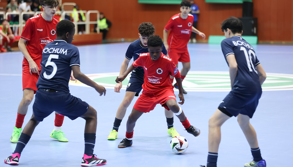 [{"clang_id":1,"value":"Futsalspiel: Vier junge Spieler, zwei mit roten, zwei mit dunklen Trikots, kämpfen um den Ball in einer Sporthalle. Im Hintergrund weitere Spieler."}]