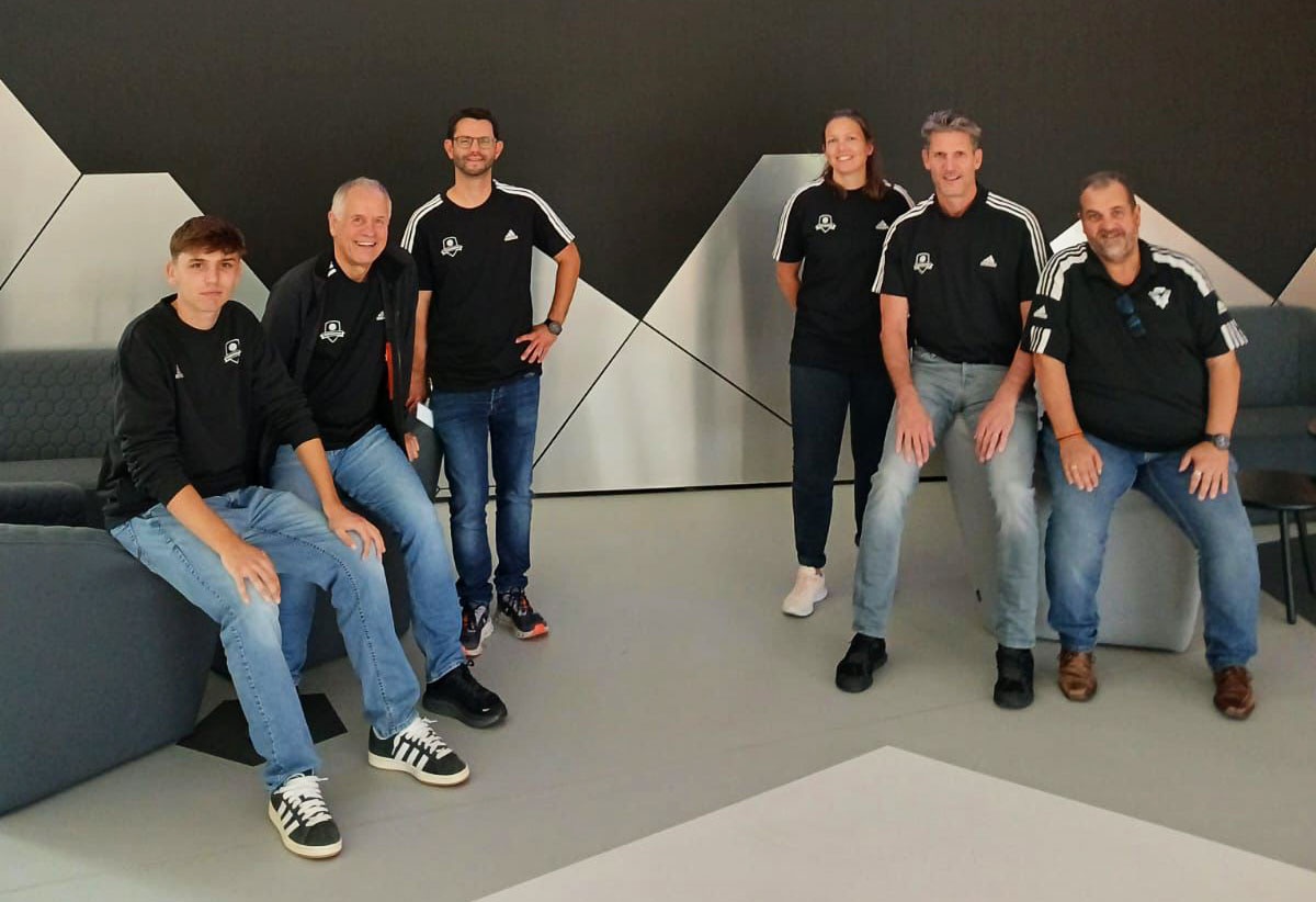 [{"clang_id":1,"value":"Sechs Personen in sportlicher Kleidung sitzen und stehen vor einer dunklen Wand mit geometrischen Formen. Alle tragen dunkle Pullover mit Logo."}]