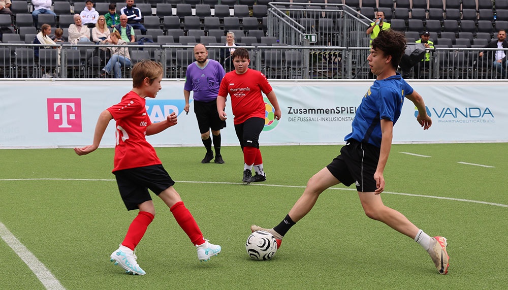 Drei Kinder spielen Fußball auf Kunstrasen. Links zwei in Rot, rechts eins in Blau. Im Hintergrund leere Zuschauertribüne und Werbung.