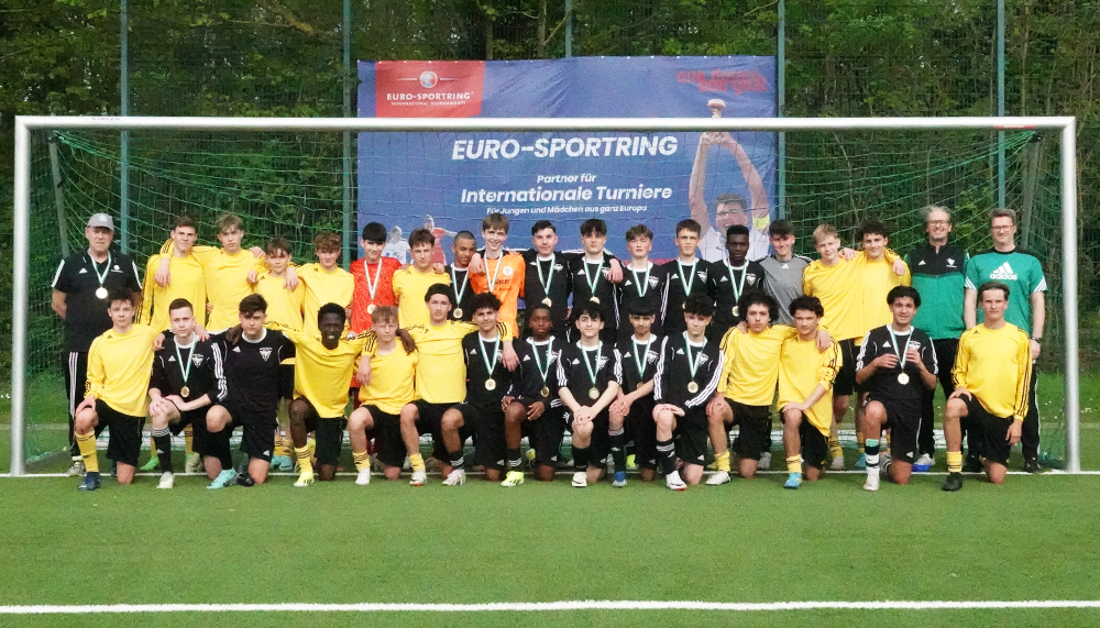 [{"clang_id":1,"value":"Fußballmannschaft mit Trainern und Betreuern posiert vor einem Fußballtor mit Werbebanner \"EURO-SPORTING Internationale Turniere\". Rasen im Hintergrund."}]