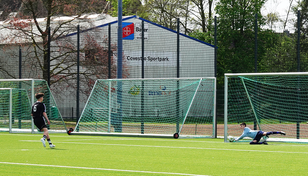 [{"clang_id":1,"value":"Fußballspieler schießt auf Tor. Im Hintergrund ein blaues Gebäude mit Schriftzug \"Covestro Sportpark\" und Bäume."}]