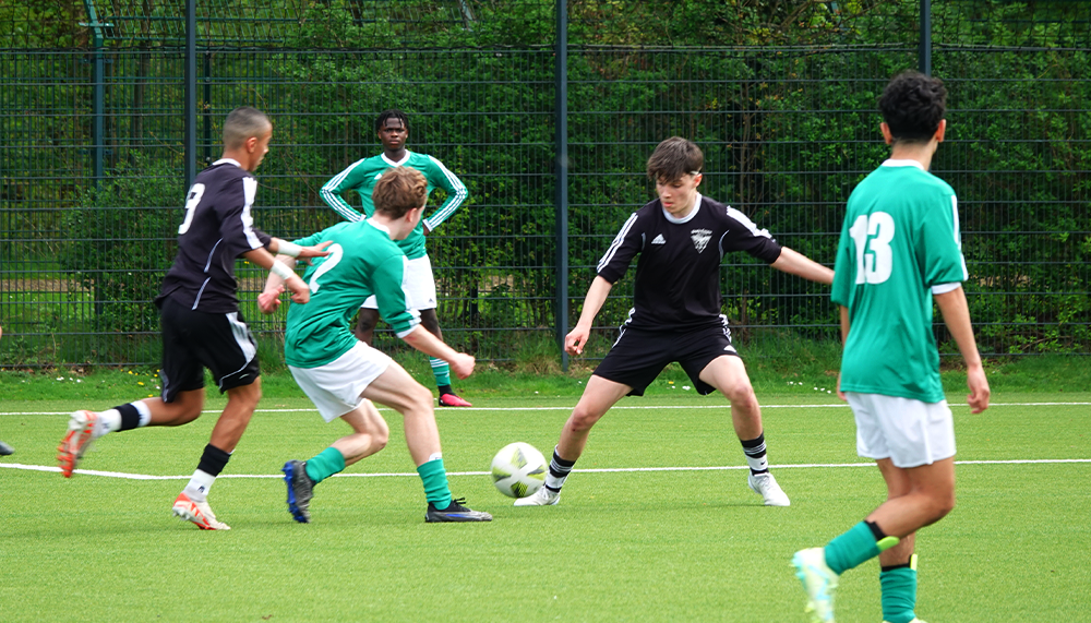 [{"clang_id":1,"value":"Fußballspiel: Vier junge Spieler auf grünem Kunstrasen, zwei in schwarzen, zwei in grünen Trikots. Ein Spieler dribbelt den Ball. Im Hintergrund ein Zaun und Bäume."}]