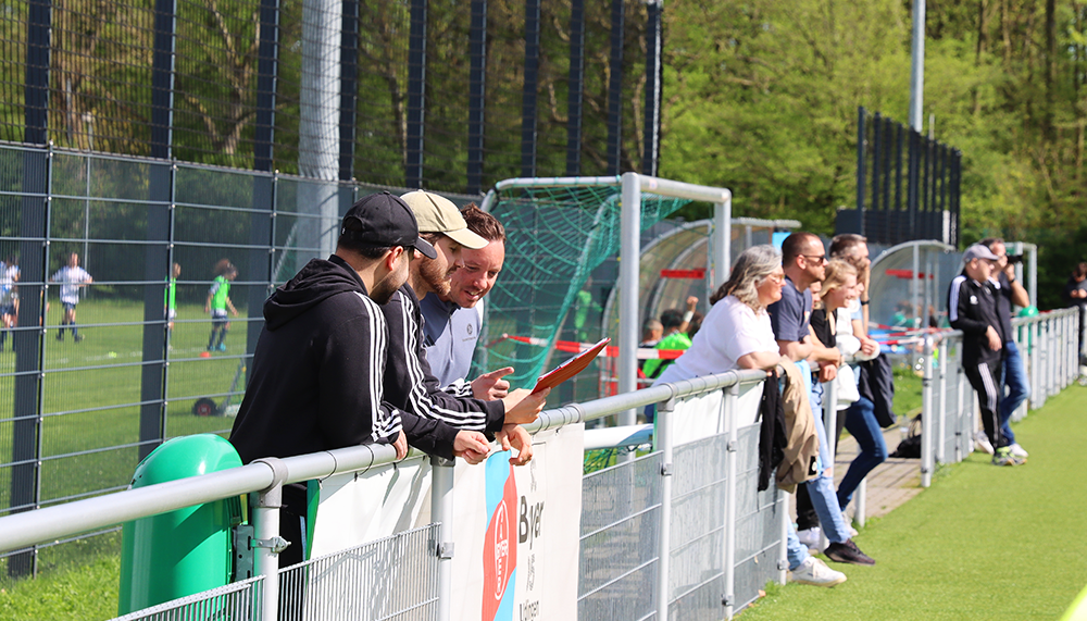 [{"clang_id":1,"value":"Fußballspiel: Zuschauer stehen an einer Absperrung und verfolgen das Spiel auf einem grünen Rasenplatz. Einige Personen schauen auf Mobiltelefone. Bäume im Hintergrund."}]