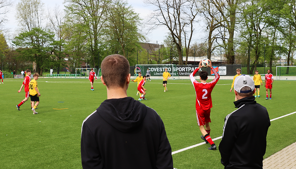 [{"clang_id":1,"value":"Fußballspiel auf Rasenplatz. Zwei Zuschauer mit dem Rücken zum Betrachter beobachten das Spiel, während ein Spieler in Rot zum Schuss ausholt. Im Hintergrund Bäume und ein Stadiondach."}]
