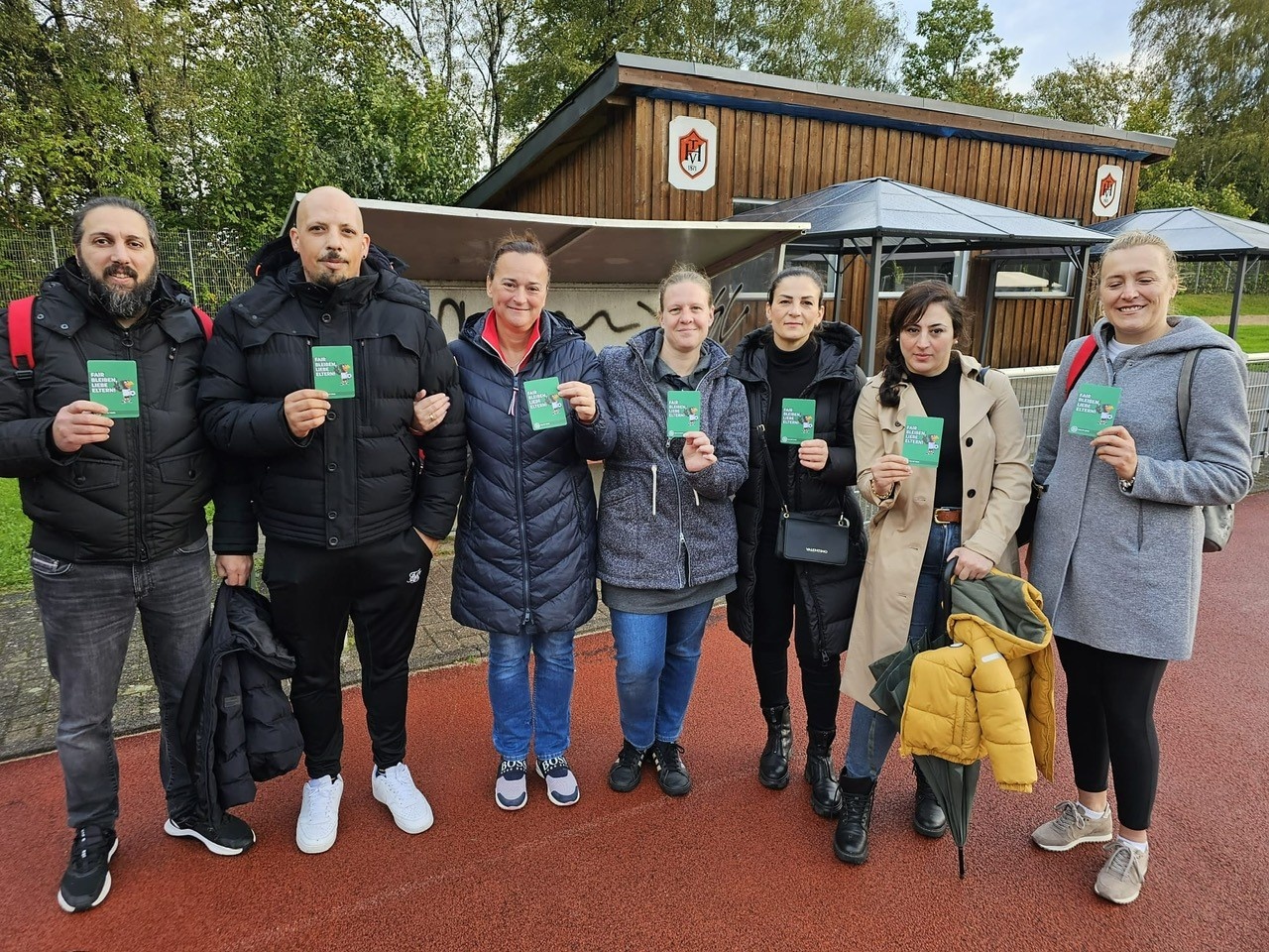 [{"clang_id":1,"value":"Gruppe von sieben Personen vor einem kleinen Gebäude mit Satteldach. Alle halten grünen Zettel hoch. Unterschiedliche Freizeitkleidung."}]