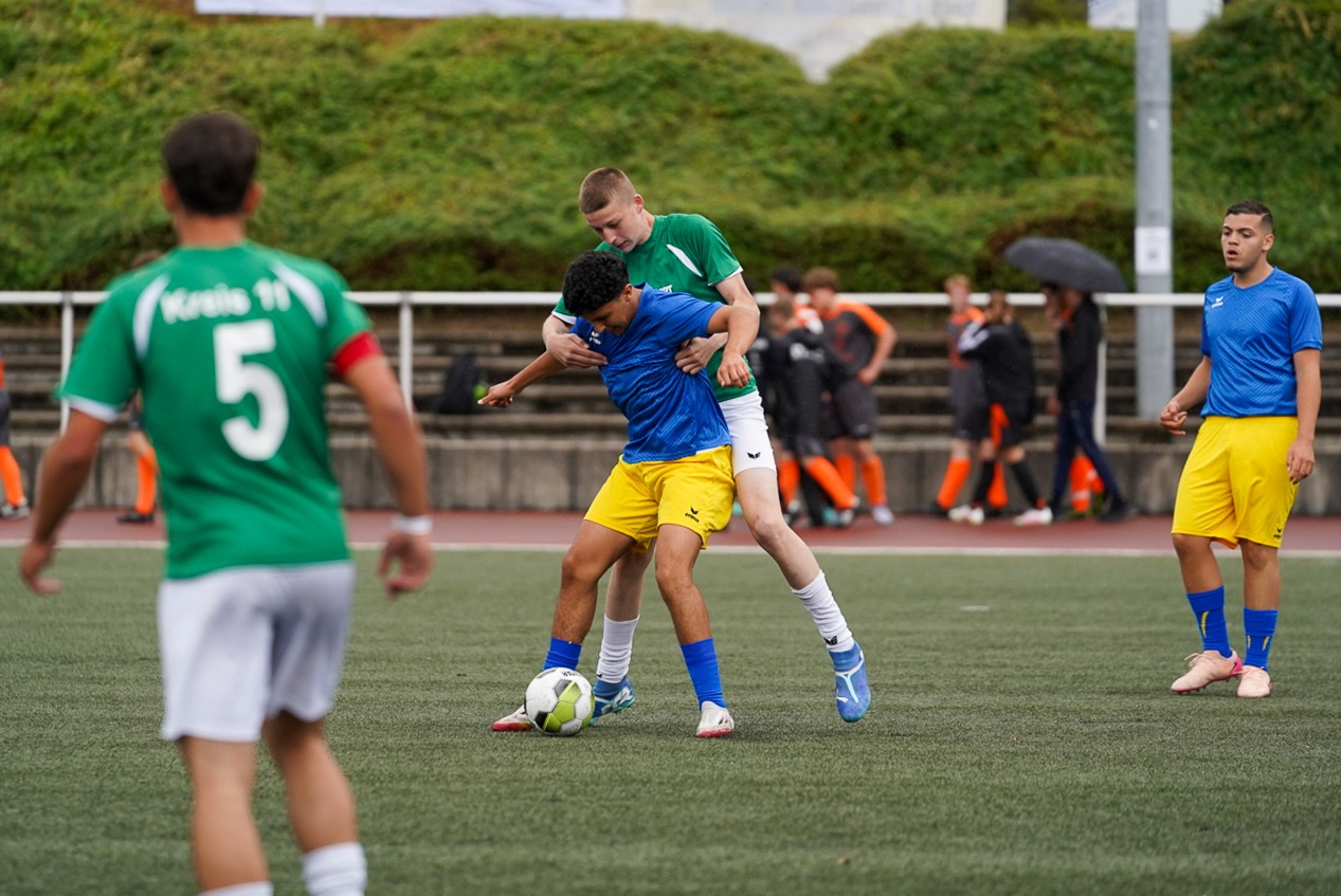 [{"clang_id":1,"value":"Fußballspiel. Zwei Spieler kämpfen um den Ball, ein dritter steht daneben. Im Hintergrund Zuschauer und Vegetation."}]