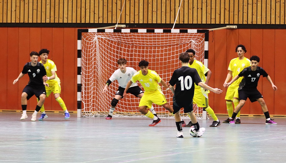 [{"clang_id":1,"value":"Futsalspiel in Sporthalle: Spieler in Schwarz und Gelb umringen Tor, Spieler mit Nummer 10 mit Ball. Holzvertäfelung im Hintergrund."}]