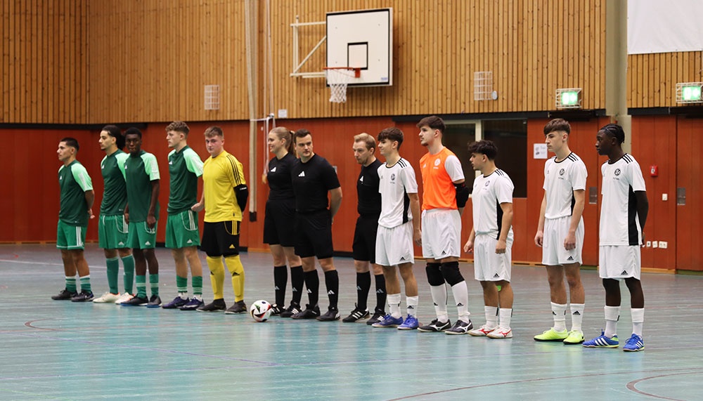 [{"clang_id":1,"value":"Futsal-Spieler in grünen, weißen und schwarzen Trikots stehen sich gegenüber, dazwischen ein Schiedsrichter. Im Hintergrund Basketballkorb und Holzwand."}]