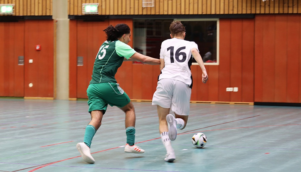[{"clang_id":1,"value":"Zwei Fußballspieler in Sporthalle. Spieler mit Trikotnummer 16 dribbelt den Ball, wird von Spieler mit Nummer 5 verfolgt."}]