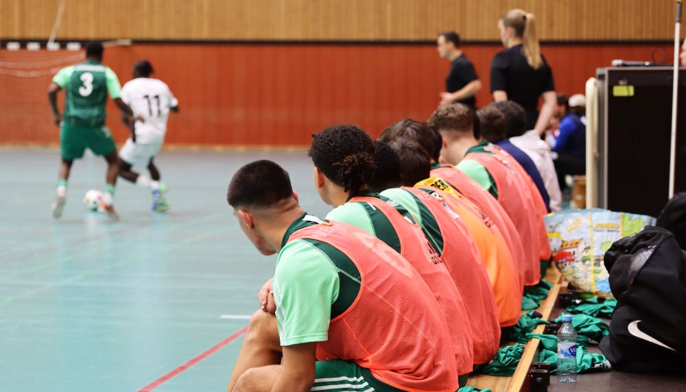 [{"clang_id":1,"value":"Sporthalle: Futsal-Spieler mit Ball im Hintergrund. Im Vordergrund sitzen Ersatzspieler auf einer Bank, alle tragen grüne Trikots und orangefarbene Westen."}]