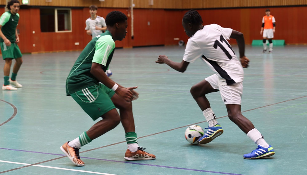 [{"clang_id":1,"value":"Zwei Fußballspieler in Hallensportarena: Einer in grünem Trikot versucht, den Ball vom Spieler mit schwarzem Trikot (Nr. 11) zu erobern. Im Hintergrund weitere Spieler."}]