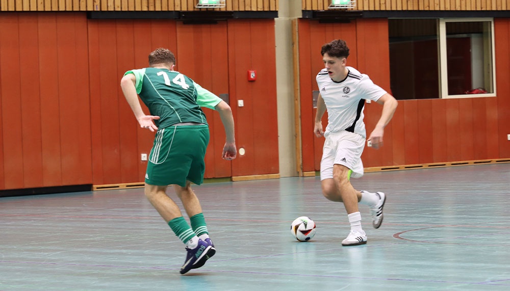 [{"clang_id":1,"value":"Zwei Fußballspieler in Sporthalle. Links Spieler mit grünem Trikot, Nummer 14. Rechts Spieler mit weiß-schwarzem Trikot, dribbelt Ball."}]