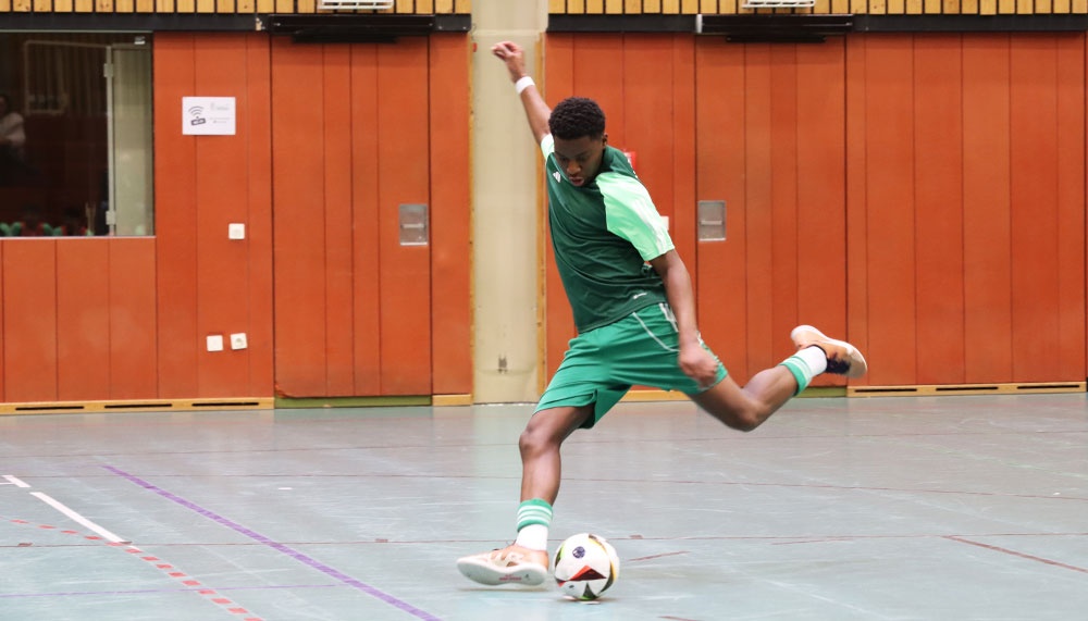 [{"clang_id":1,"value":"Fußballspieler in grüner Sportkleidung schießt in einer Sporthalle auf einen Ball. Braune Wand im Hintergrund."}]