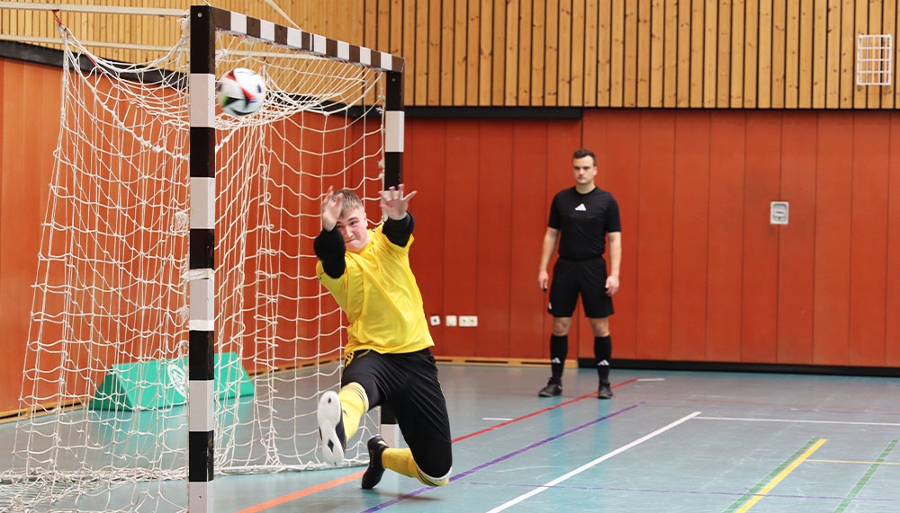 [{"clang_id":1,"value":"Handballtorwart in gelbem Trikot springt zur Ballabwehr. Im Hintergrund ein Mann und eine orangefarbene Wand. Sporthalle."}]