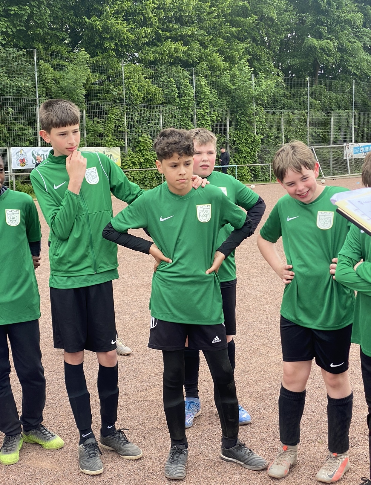 [{"clang_id":1,"value":"Fünf Jungen im Fußballtrikot (grün, schwarz) stehen auf einem Sportplatz. Ein Junge hält nachdenklich den Kopf, ein anderer lächelt breit. Im Hintergrund ein Zaun und Bäume."}]