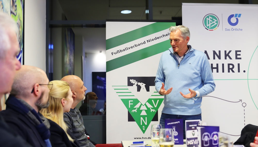 [{"clang_id":1,"value":"Mann spricht vor Publikum an einem Tisch mit Logo \"FVN\". Im Hintergrund ein Banner mit Fußballverein-Schriftzug und Werbeaufdruck."}]